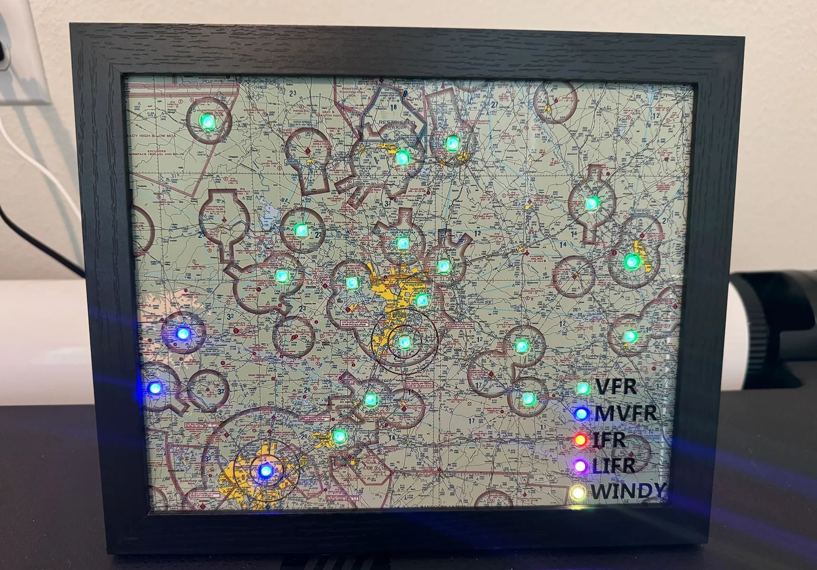 A framed sectional map on a desk with green and blue airport lights and a lit legend for VFR, MVFR, IFR, LIFR and Windy